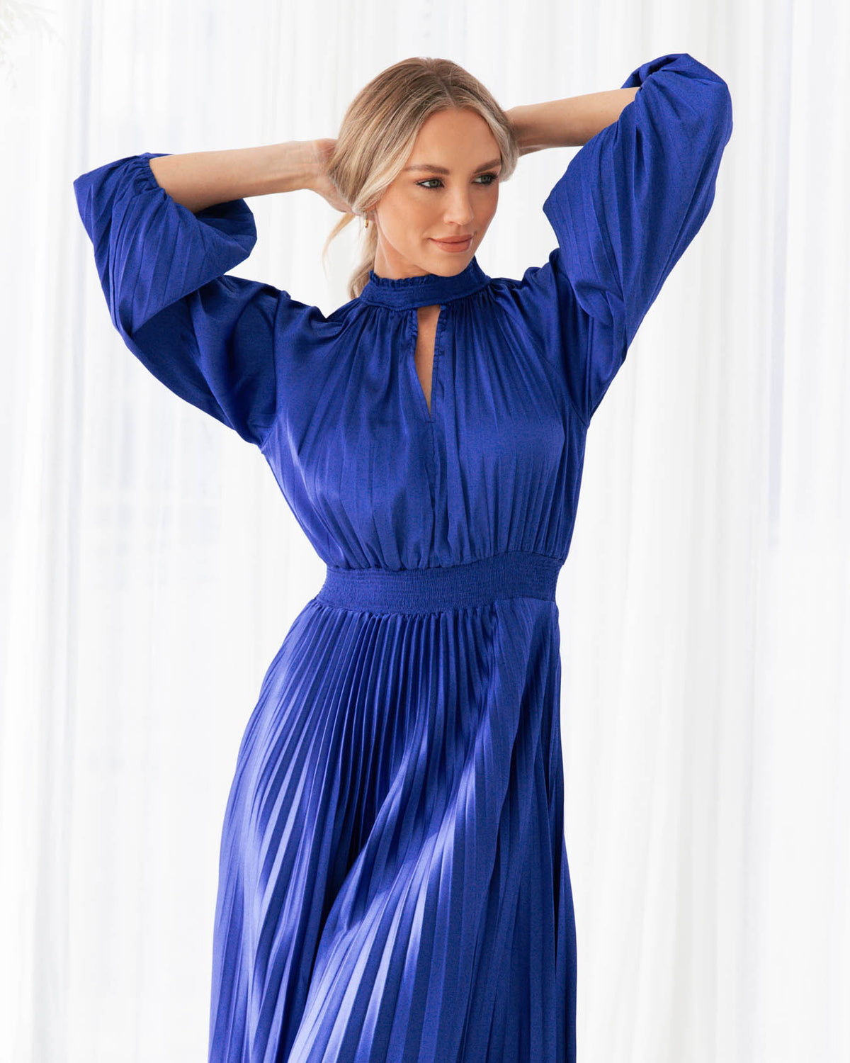 A model is wearing a blue pleated keyhole midi dress with balloon sleeves from the Ebby and I collection designed by Global Fashion House.