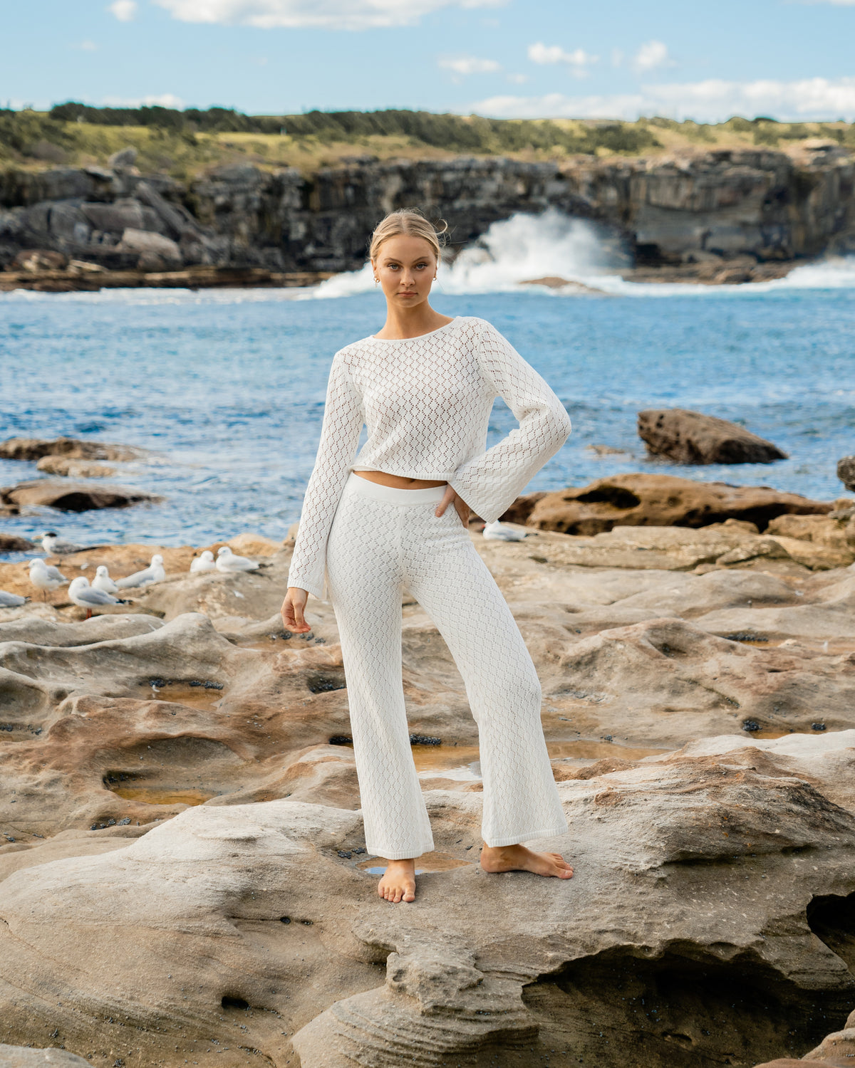 A model is at the beach wearing a white long-sleeve crochet top and white high waist crochet pants from the Paper Heart collection designed by Global Fashion House.
