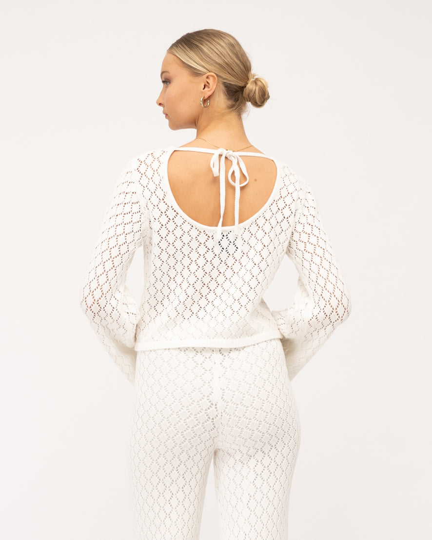 A model is at the beach wearing a white long-sleeve crochet top and pants from the Paper Heart collection designed by Global Fashion House.