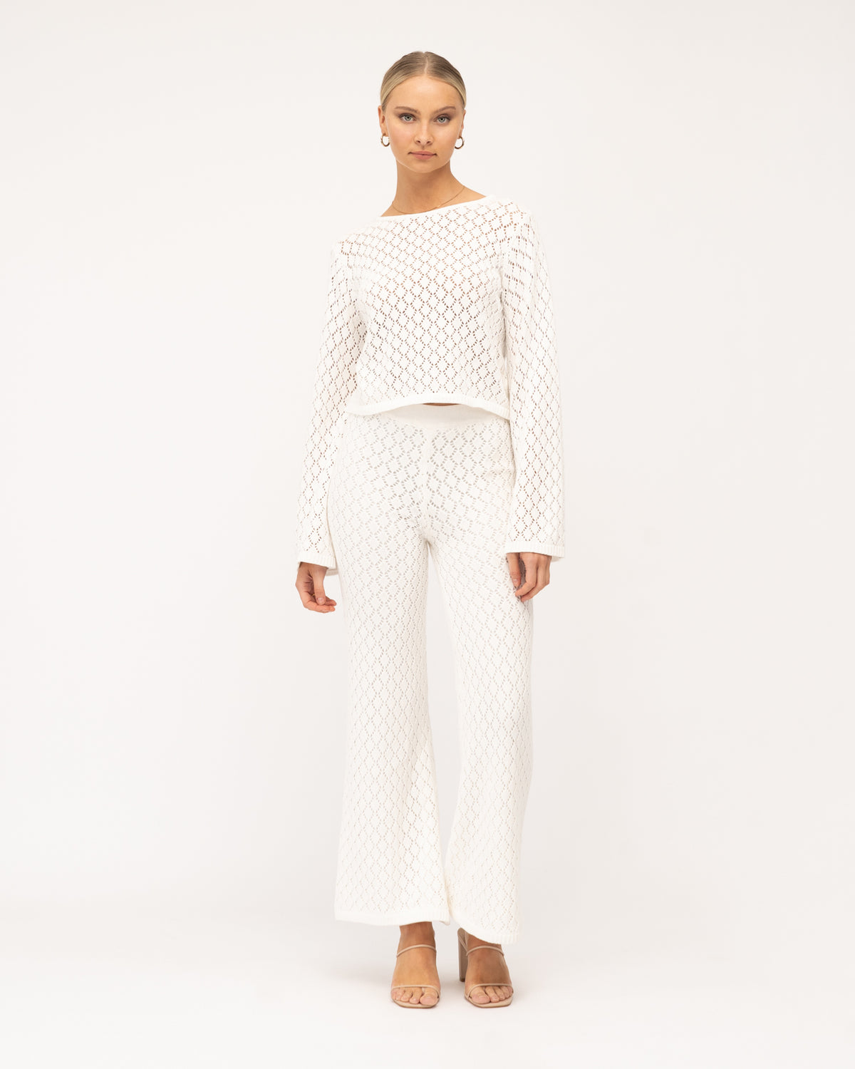 A model is at the beach wearing a white long-sleeve crochet top and pants from the Paper Heart collection designed by Global Fashion House.