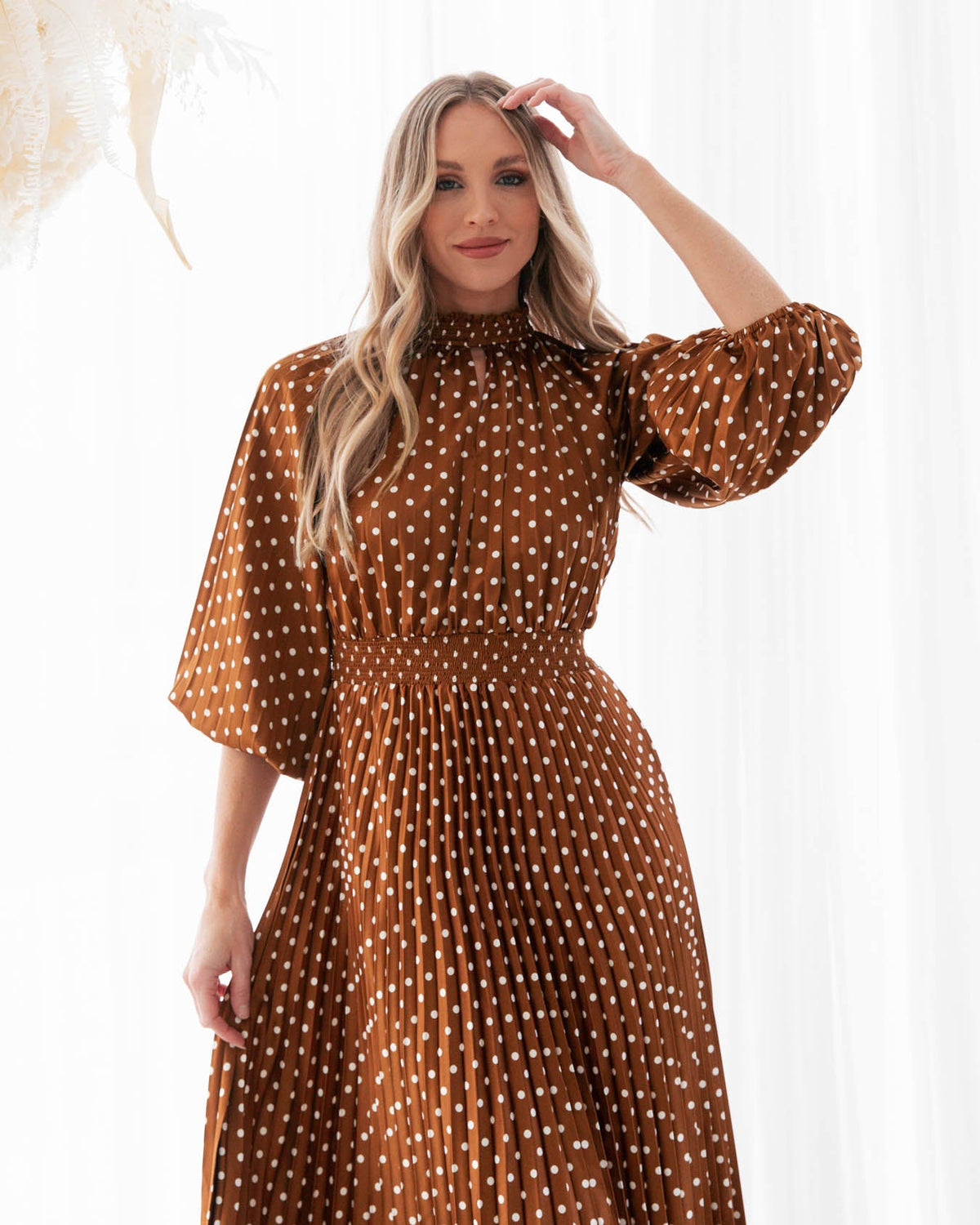 A model is wearing a rust polka dot pleated keyhole midi dress with balloon sleeves featuring elastic cuffs from the Ebby and I collection designed by Global Fashion House.