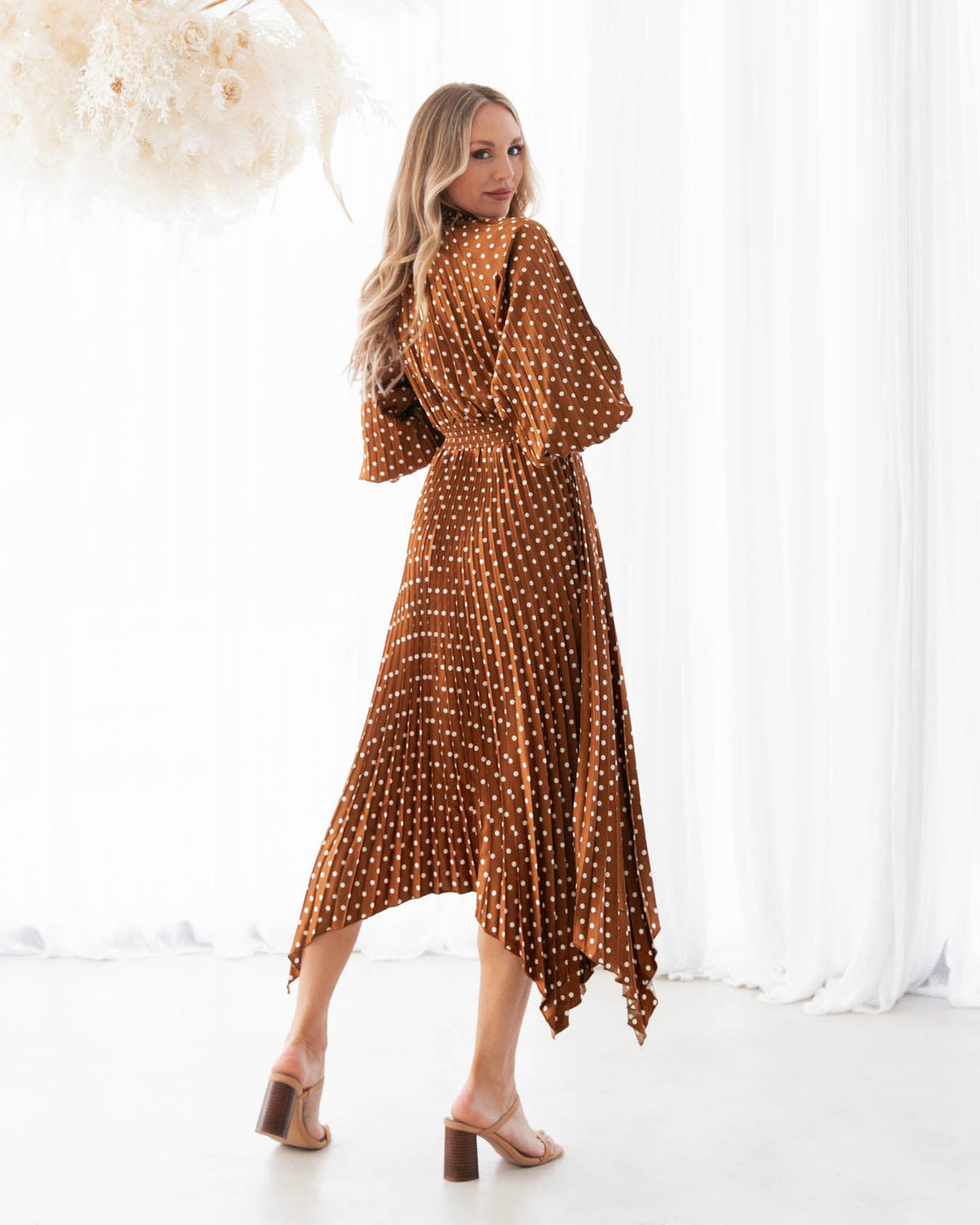 A model is wearing a rust polka dot pleated keyhole midi dress with balloon sleeves featuring elastic cuffs from the Ebby and I collection designed by Global Fashion House.